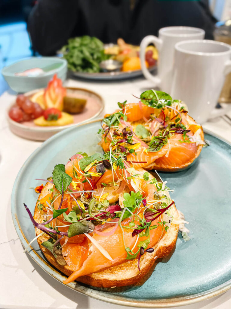 Blue plate with open-faced smoked salmon bagel, pickled fennel, and microgreens beside steaming coffee mugs.