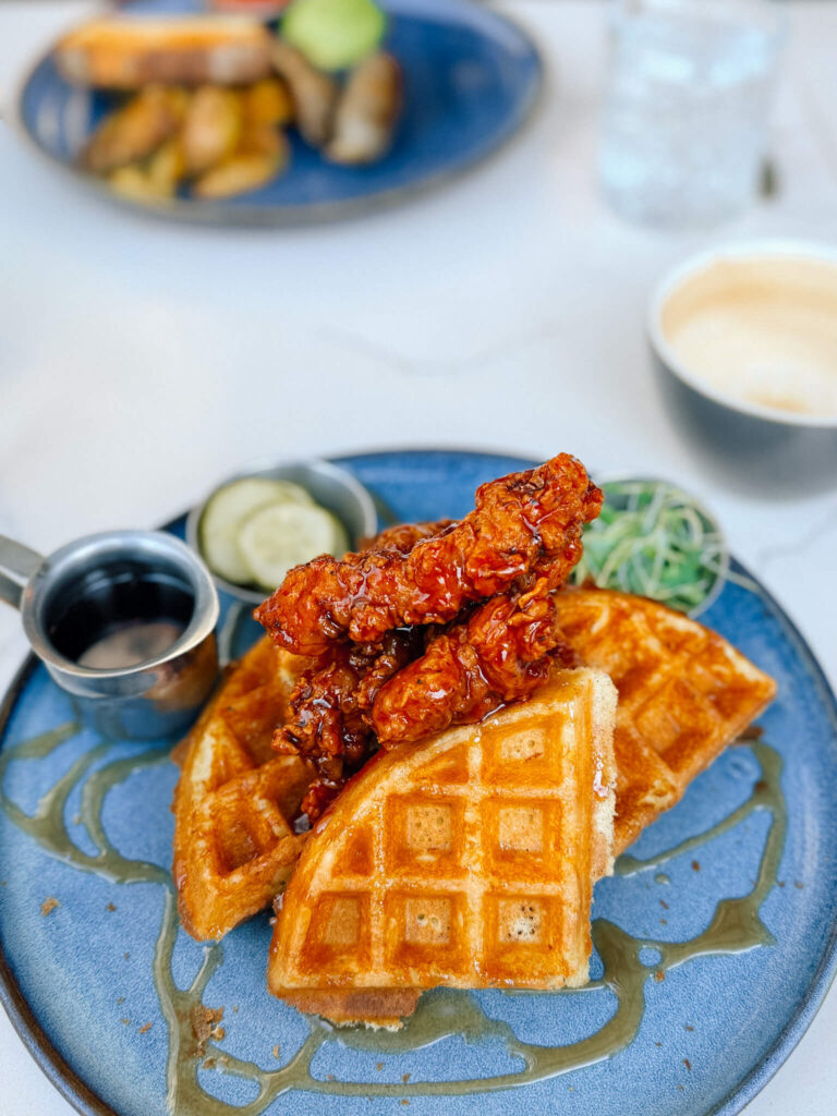 A tall, layered stack of golden waffles and crunchy sweet and spicy chicken covered in warm salted caramel and rich Québec maple syrup, usually topped with micro arugula and tangy pickles, seen here on the side.