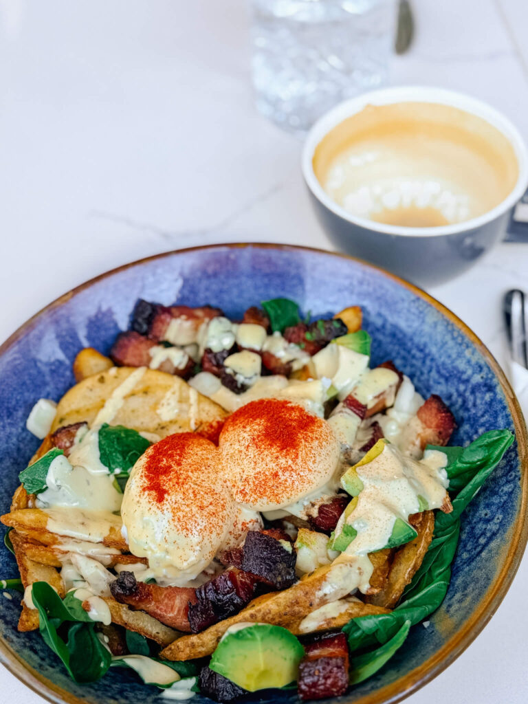 A gourmet breakfast poutine topped with soft poached eggs, a dusting of paprika, silky hollandaise, golden herb-roasted potatoes, spinach, artisan cheese curds, and thick-cut bacon lardons.