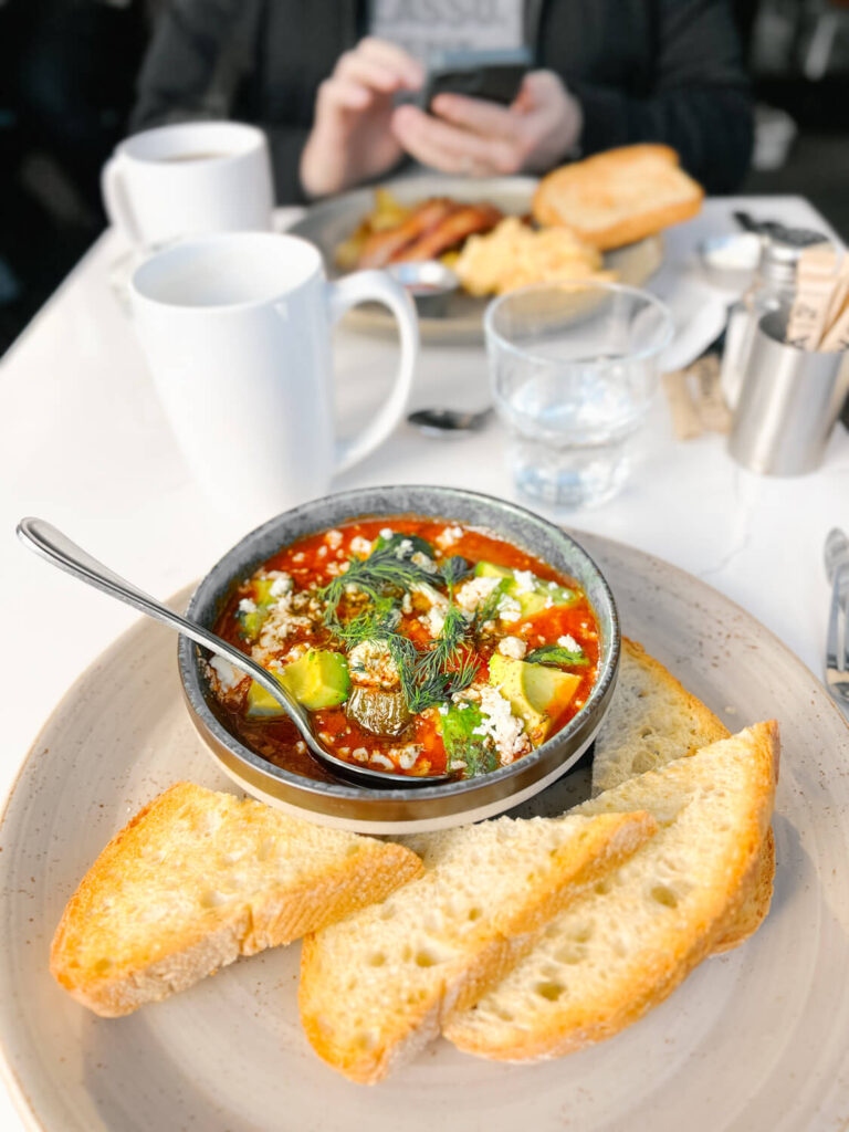 A bowl filled to the brim with stewed tomato shakshuka topped with feta cheese, avocado, and fresh dill. There is also perfectly toasted sourdough on the side, and steaming cups of coffee.